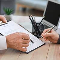 A person at a desk with a clipboard showing another person where to sign a document with their pen