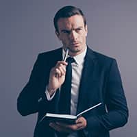 A person standing in a suit holding a book looking up in a thinking gesture