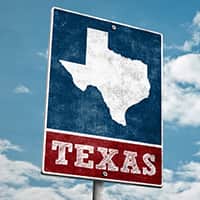 A road sign that has a picture of the state of Texas with the word Texas below. Behind the sign is a blue sky with clouds