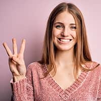 Person smiling in a pink sweater holding three fingers up on their right hand